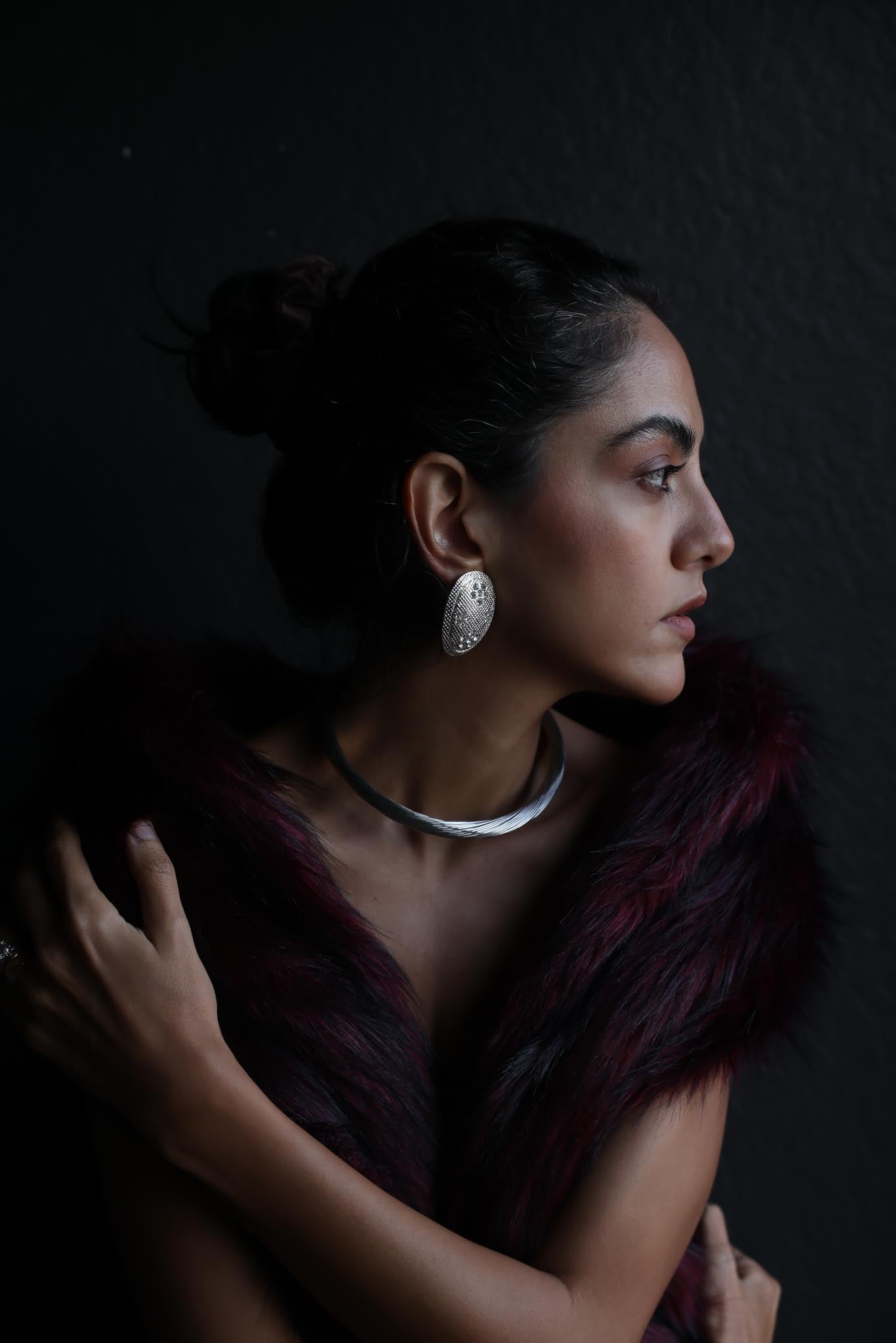 Side profile of a woman wearing a vintage silver-tone collar necklace with a twisted design and oval textured clip-on earrings, styled with a burgundy faux fur wrap against a dark background.