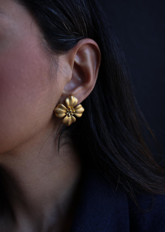 Close-up of a woman’s ear adorned with a matte gold floral pierced earring featuring delicate petal details and a textured center. The earring has a timeless and elegant design.